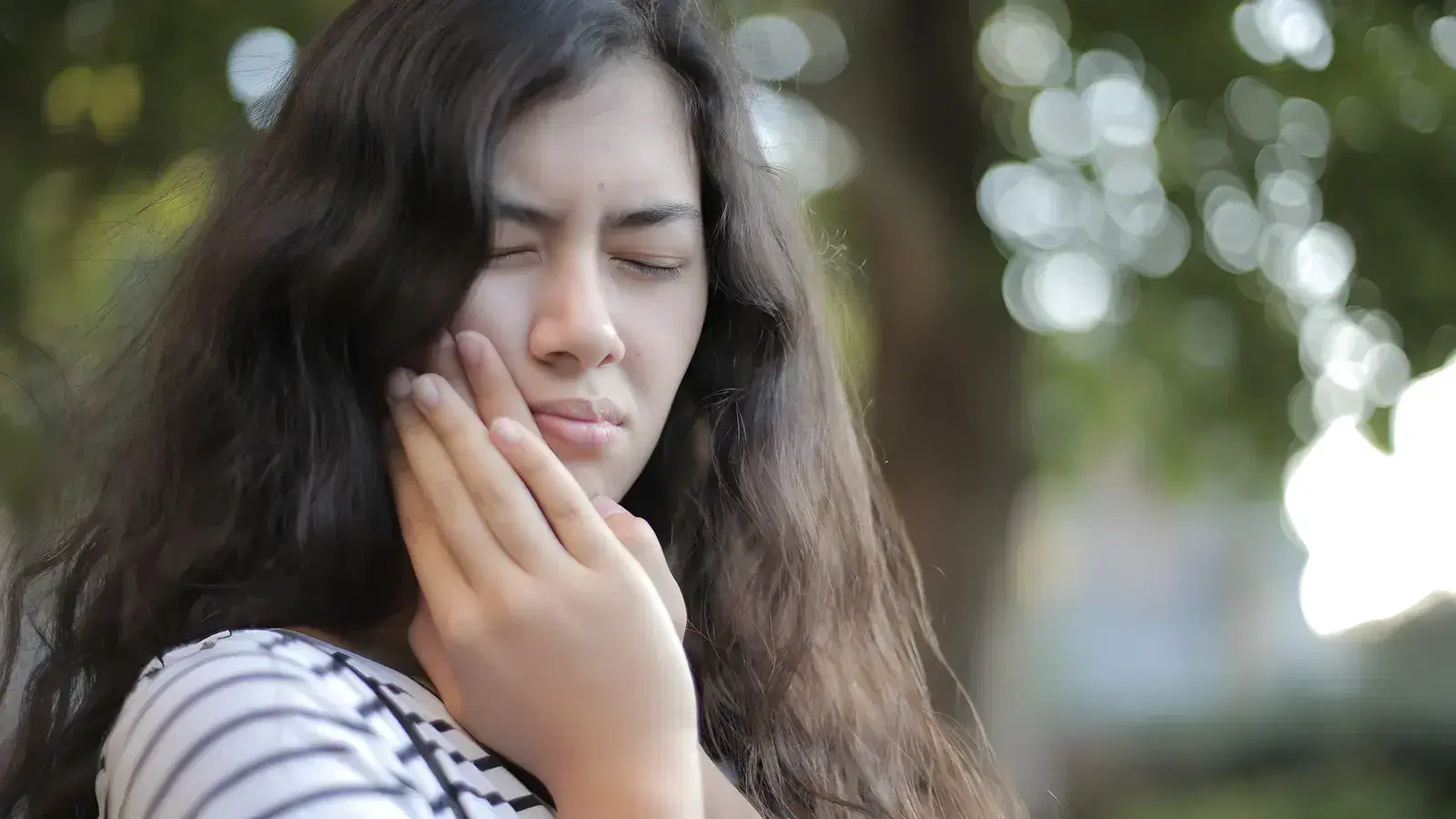 A young woman outdoors in dappled daylight, eyes shut and hand pressed to her jaw as a dental abscess throbs