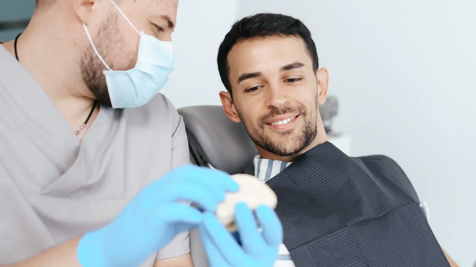 A dentist showing a smiling patient in the chair a tooth model while explaining crown treatment